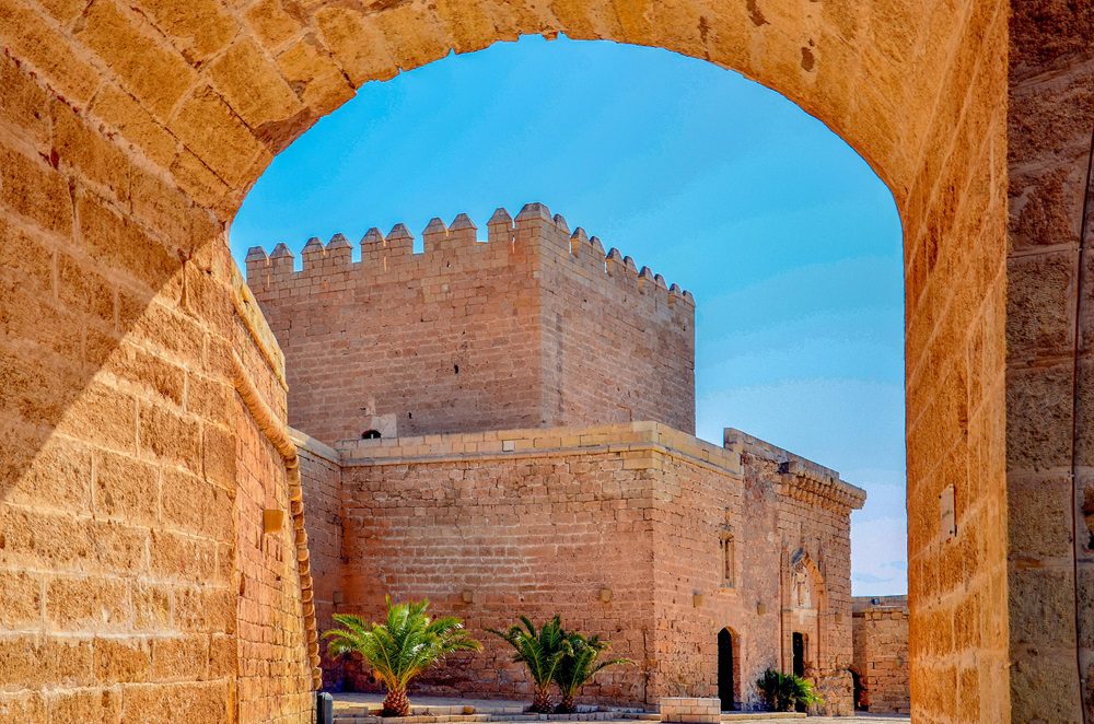 Arco y muro histórico con torre de piedra en Almería, España, con cielo azul despejado.