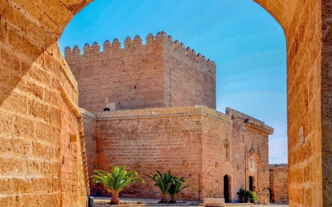 Arco y muro histórico con torre de piedra en Almería, España, con cielo azul despejado.
