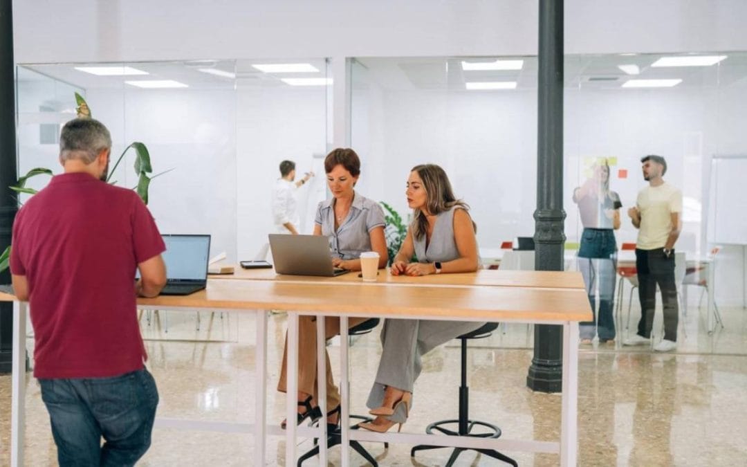 Personas trabajando y conversando en un espacio de coworking moderno y luminoso con mesas compartidas y ordenadores portátiles.